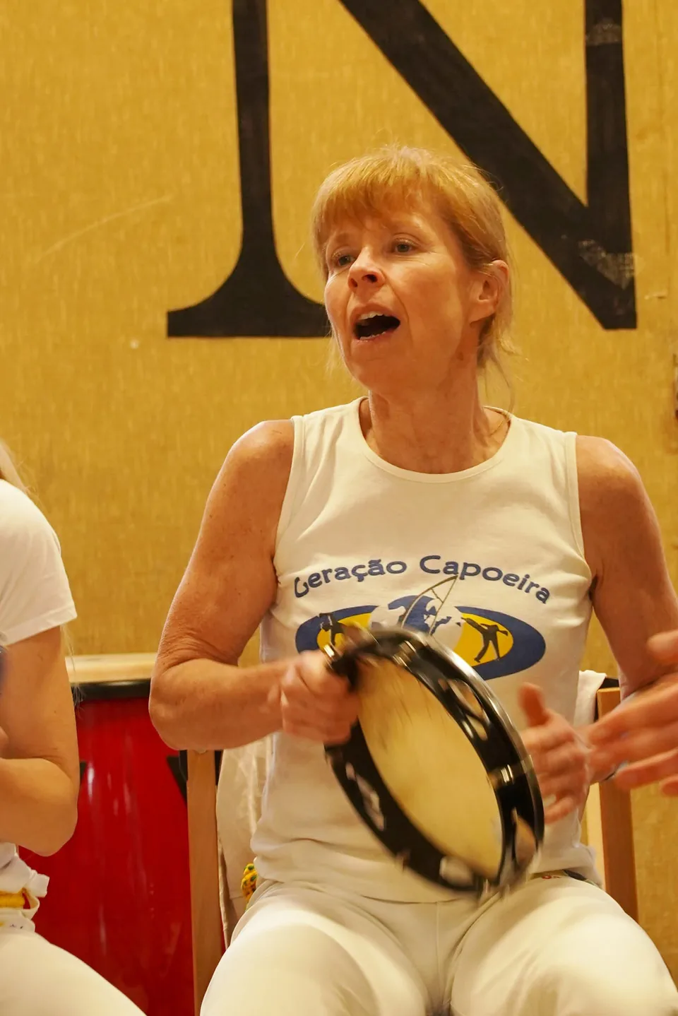 Une capoeiriste chante en jouant du pandeiro, vêtue du débardeur de l’association.