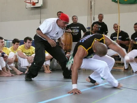 Due capoeiristi giocano vicino al suolo davanti ai partecipanti seduti e ai musicisti.