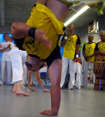 Un capoeirista con camiseta amarilla mantiene el equilibrio sobre una mano en la roda.